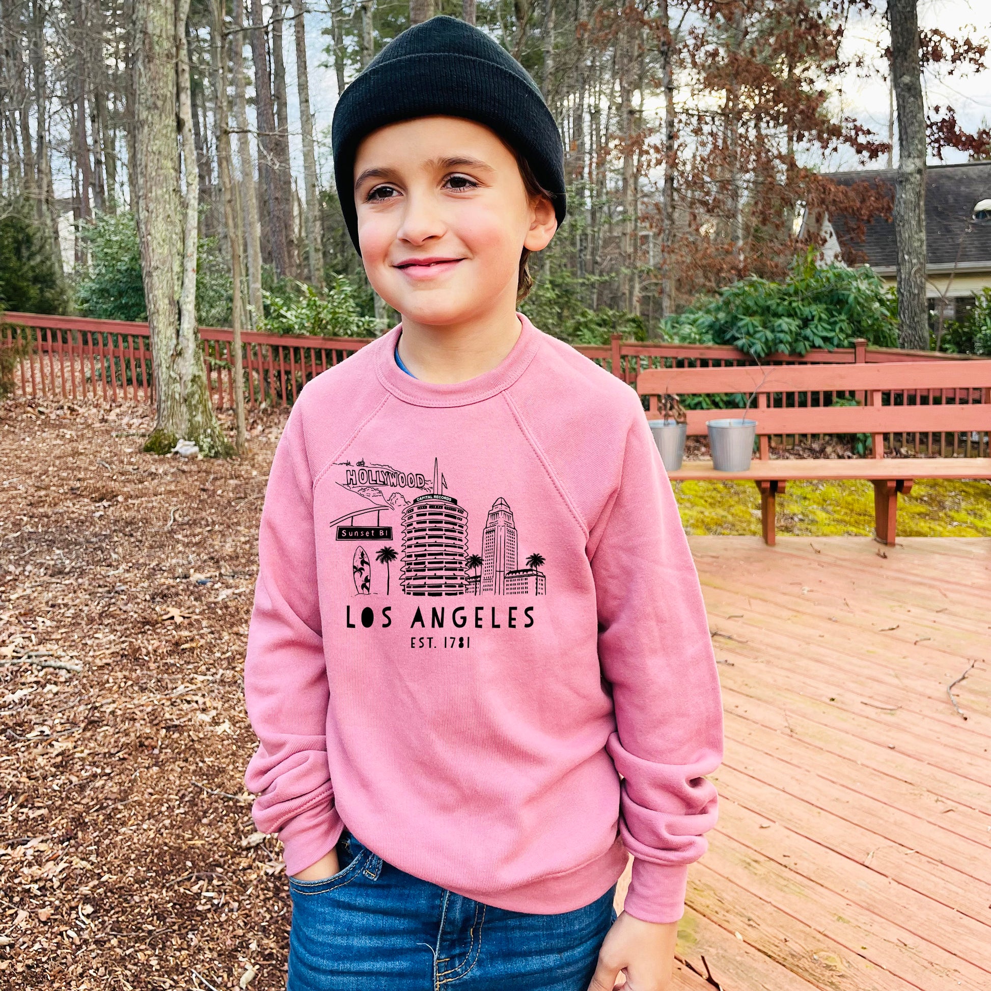 a young boy standing on a deck wearing a pink los angeles sweatshirt