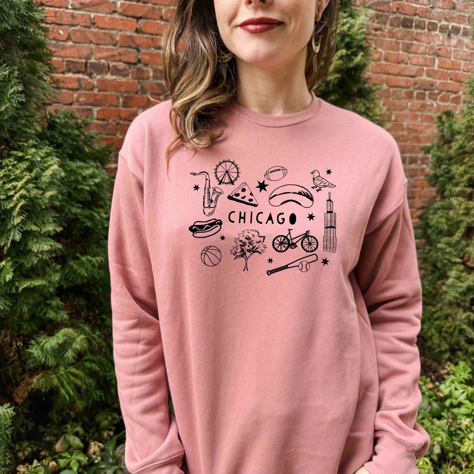 a woman wearing a pink chicago sweatshirt stands in front of a brick wall